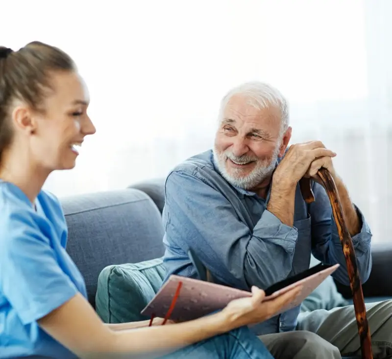 Elderly man sitting on a sofa holding a walking stick, with a carer beside him