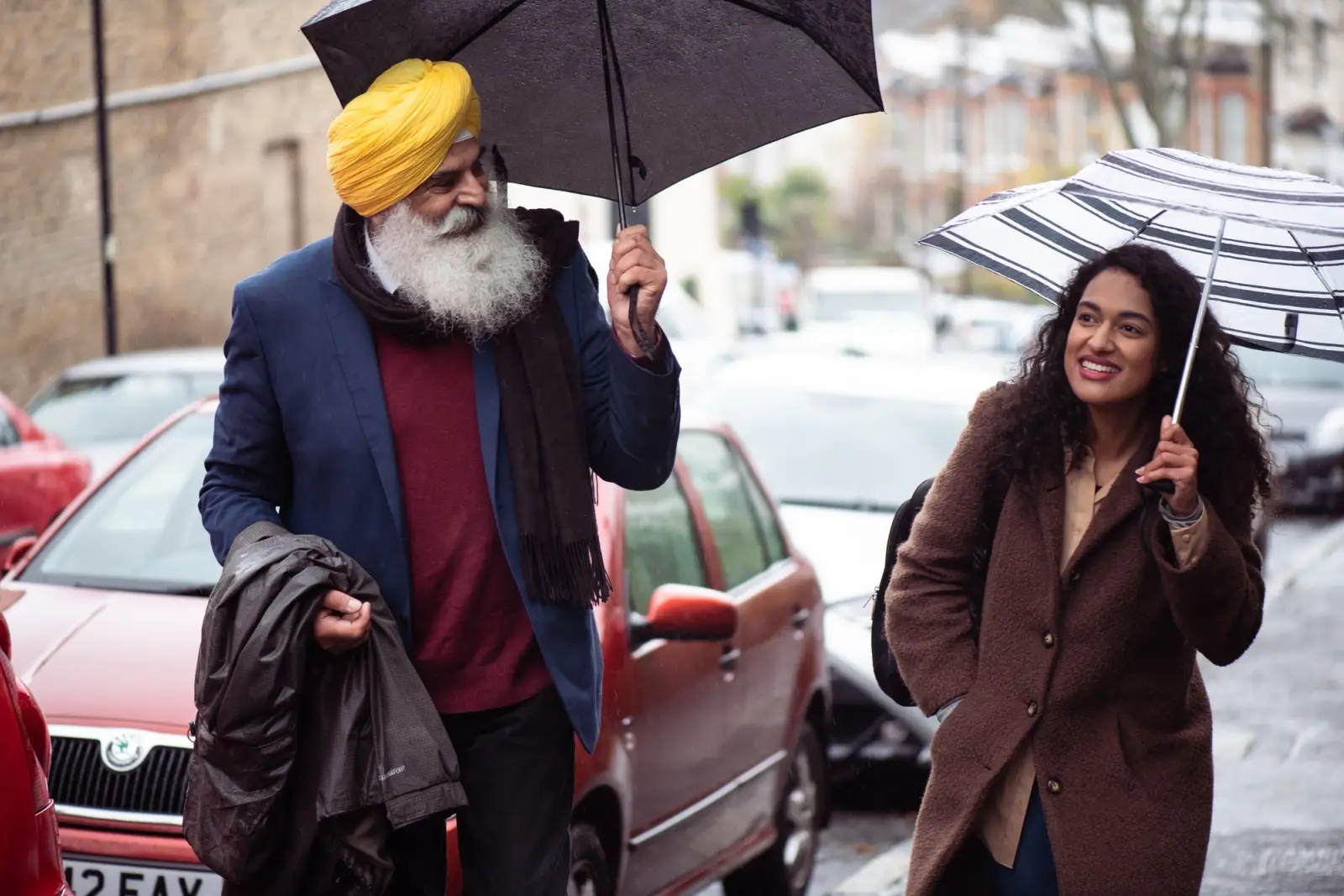 An elderly man taking a walk with his adult daughter in the street in the rain, both are using an umbrella.