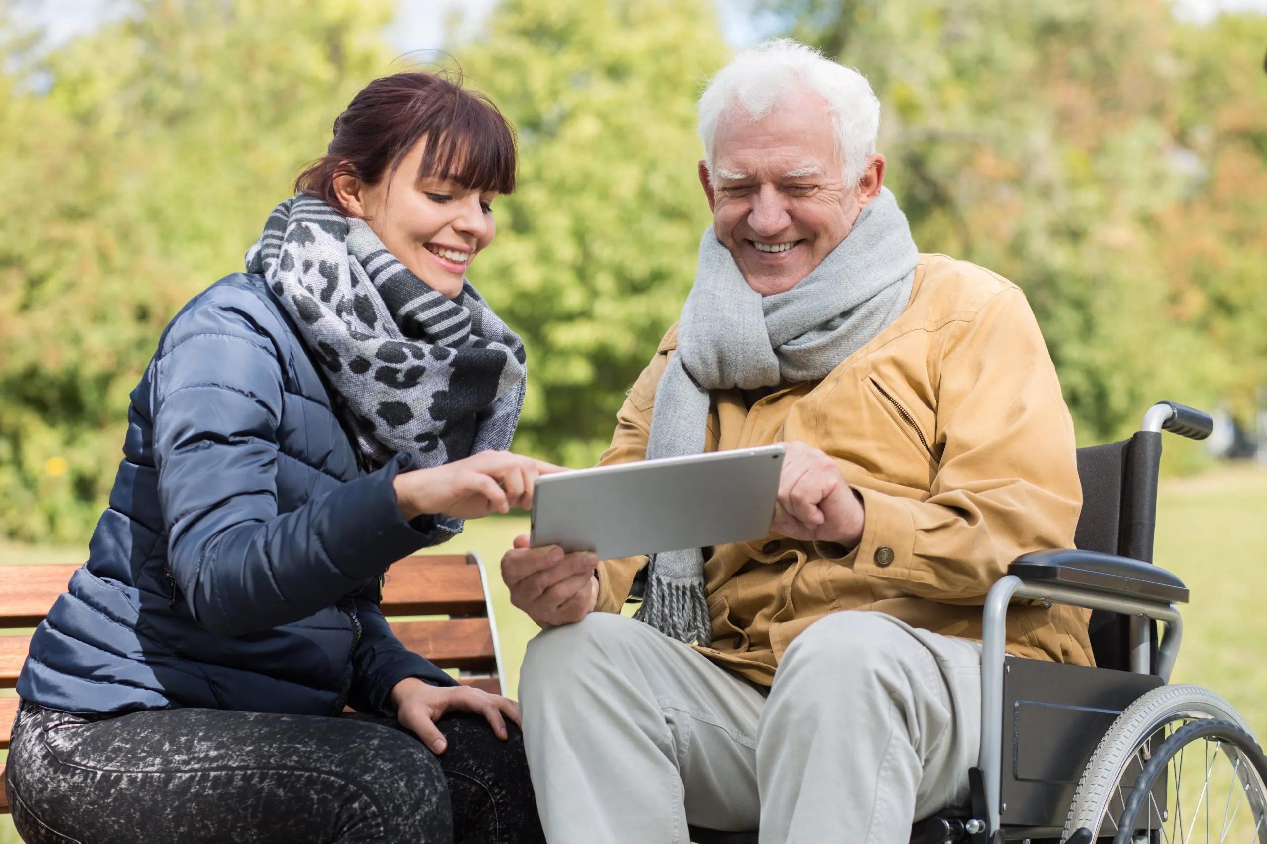 A carer sits on a park bench next to an elderly man in a wheelchair