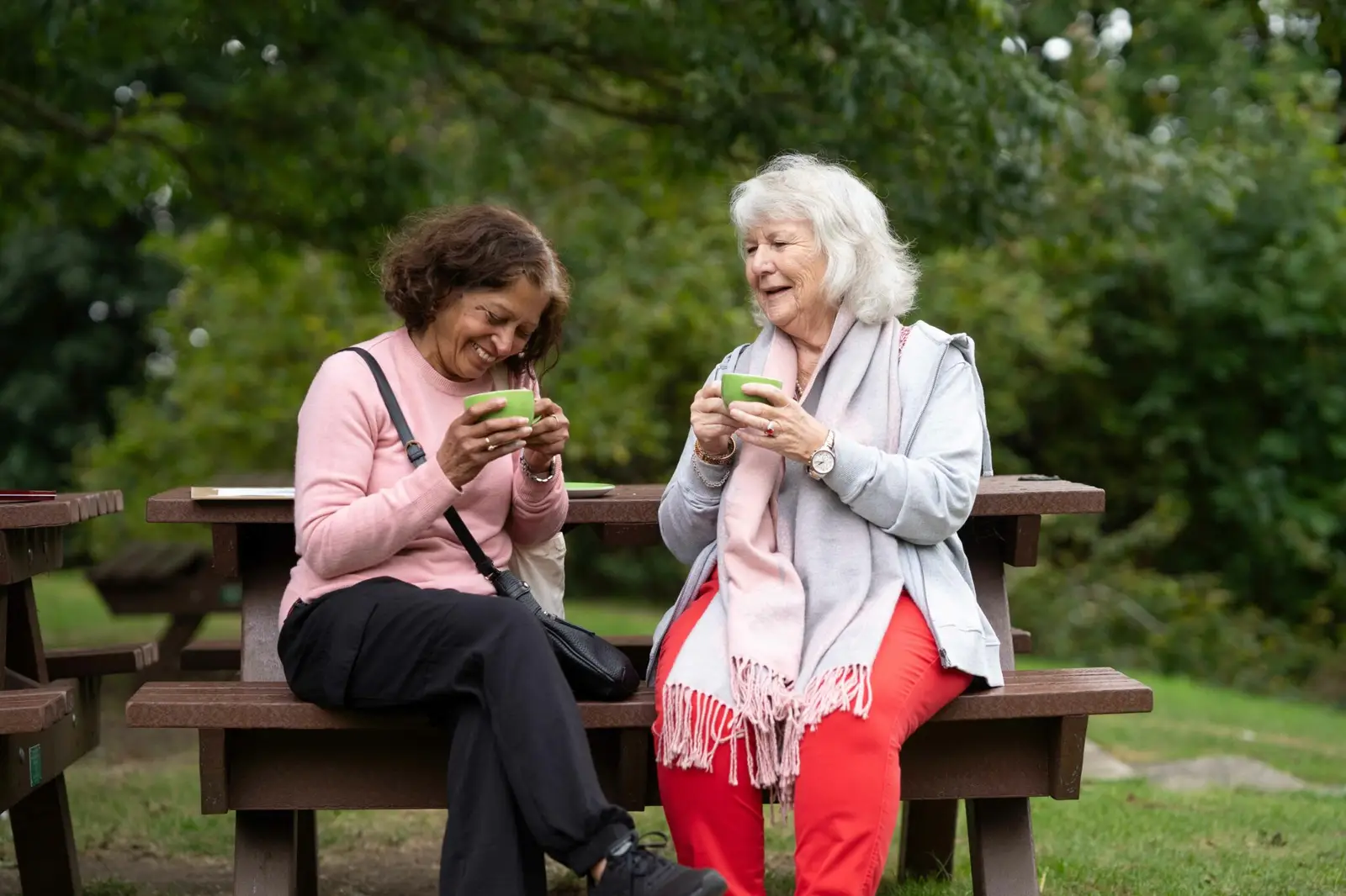 Elderly woman sits on a bench with her carer drinking a cup of tea