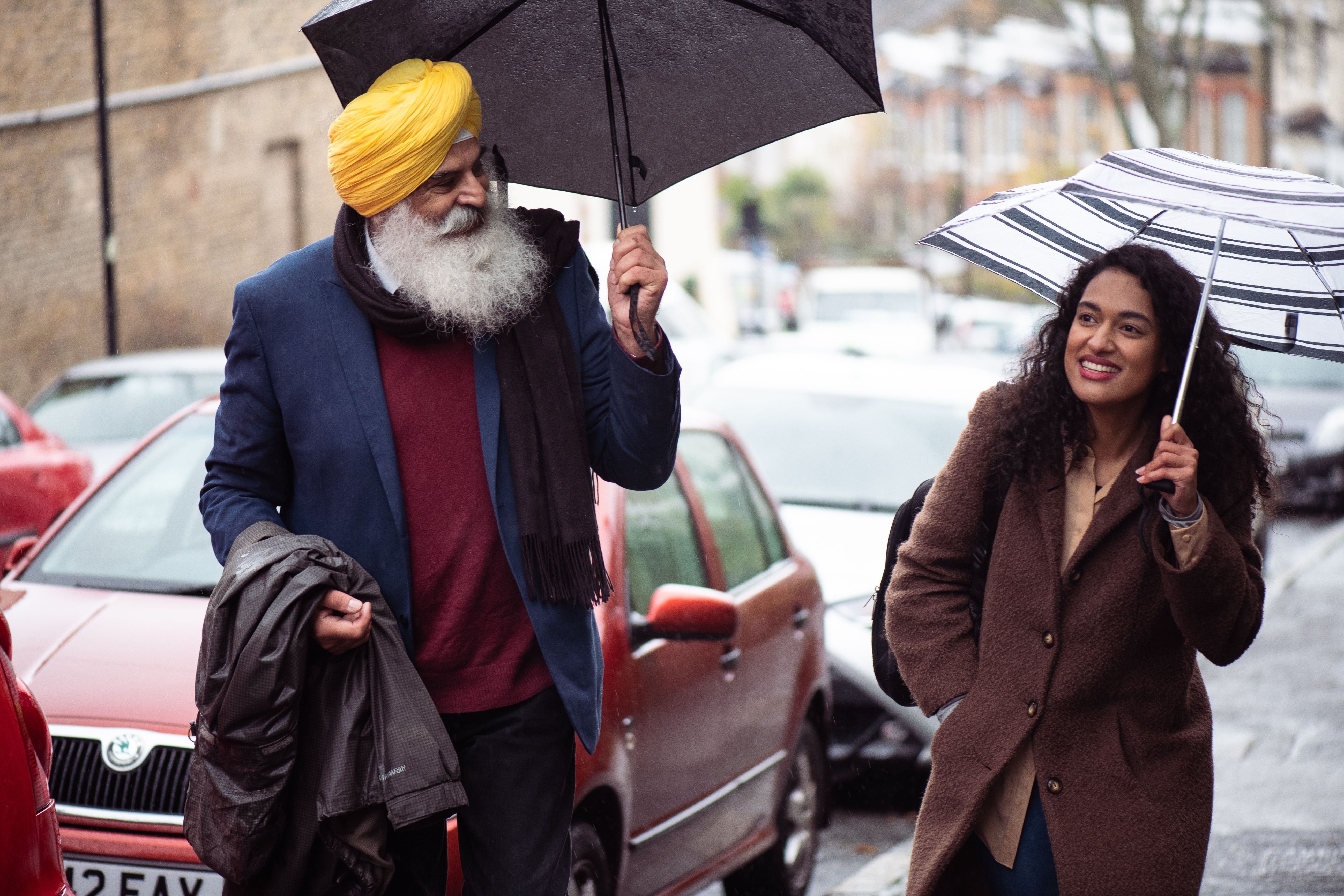 An elderly man taking a walk with his adult daughter in the street in the rain, both are using an umbrella.