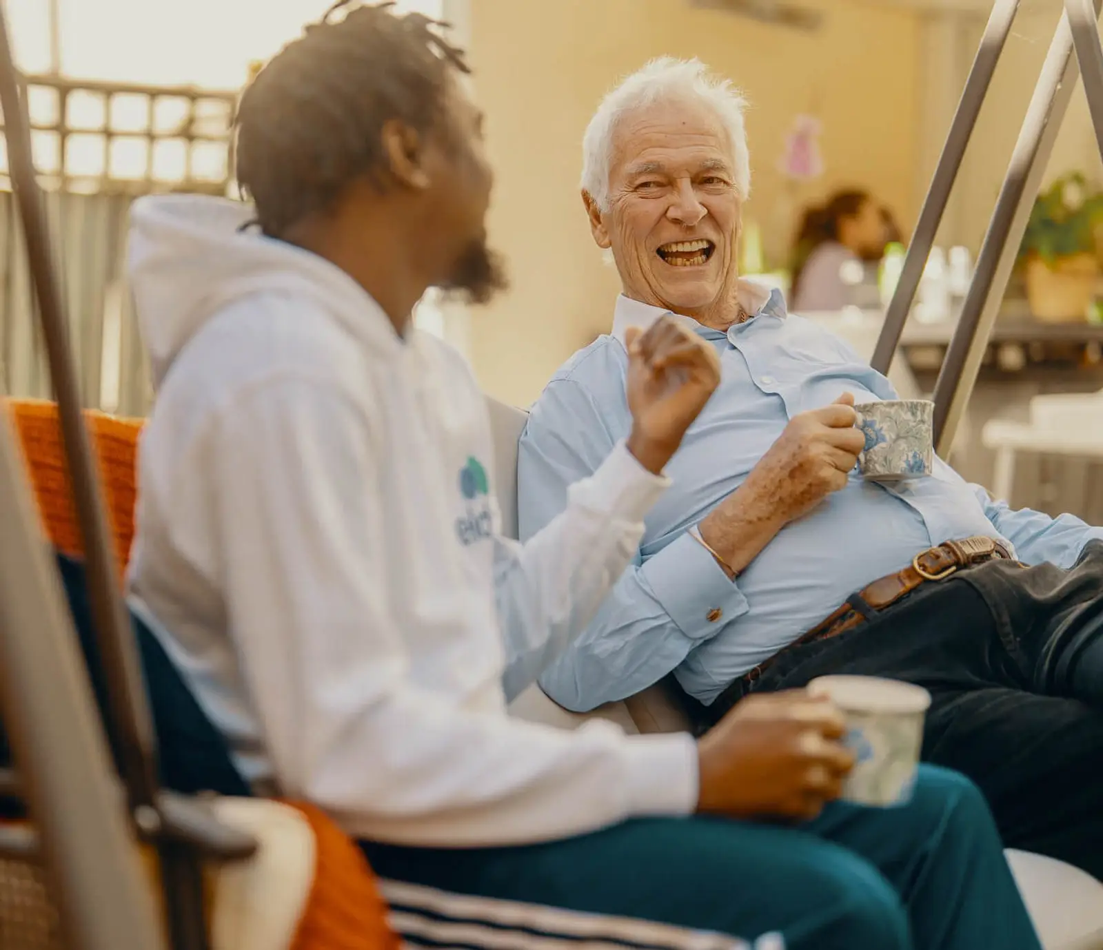 An Elder live-in carer enjoys a cup of tea with his client on a garden swing seat.