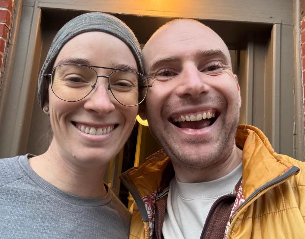 A smiling woman with glasses and a headscarf stands next to a smiling man in a yellow jacket, both posing for a selfie in front of a doorway with warm lighting. The background features a textured wall that adds character to the scene.