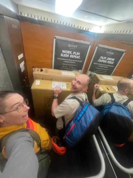 Two individuals are crammed into a small elevator, each holding onto a large cardboard box while wearing backpacks. They both smile at the camera, surrounded by the elevator’s wooden paneling and a promotional poster for a hotel.