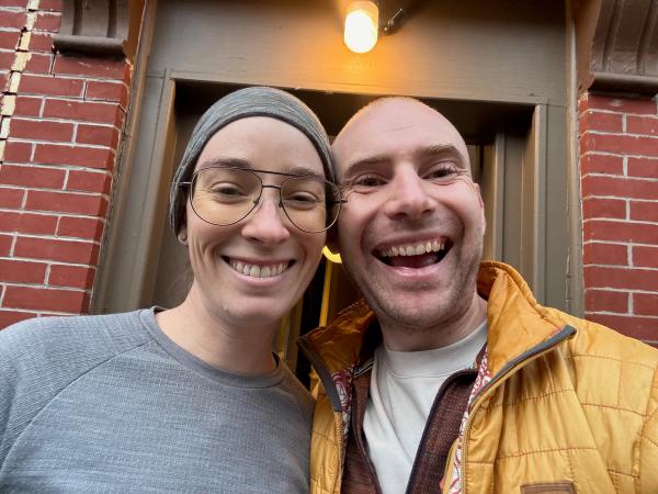 A smiling woman with glasses and a headscarf stands next to a smiling man in a yellow jacket, both posing for a selfie in front of a doorway with warm lighting. The background features a textured wall that adds character to the scene.