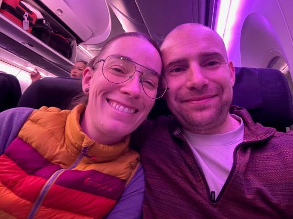 A smiling couple sits close together on an airplane, with the woman wearing large glasses and a colorful puffy vest in shades of orange and purple. The cabin is illuminated with soft purple lighting, and a few passengers can be seen in the background.