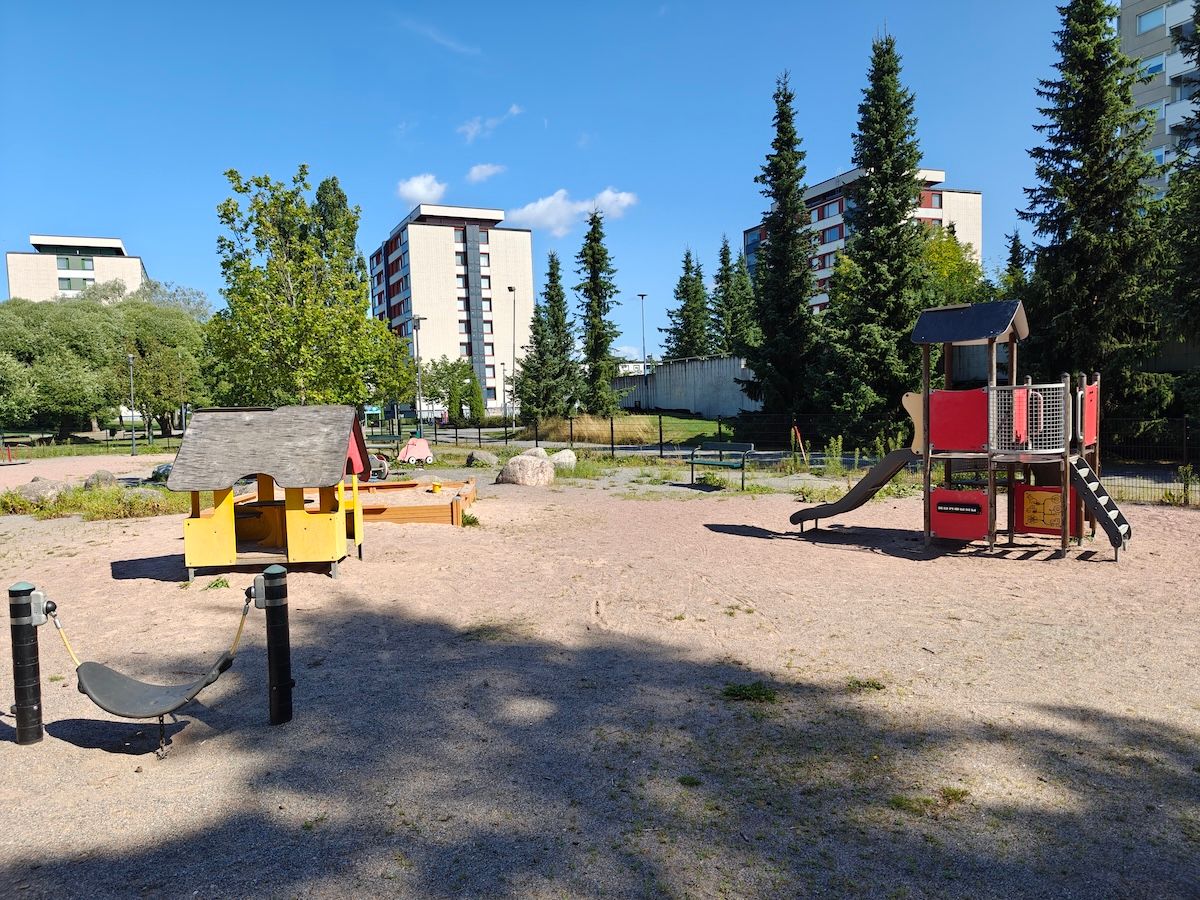 Leikkimökki, hiekkalaatikko ja pienemmille lapsille sopiva kiipeilyteline liukumäellä. Taustalla jousivälineitä.