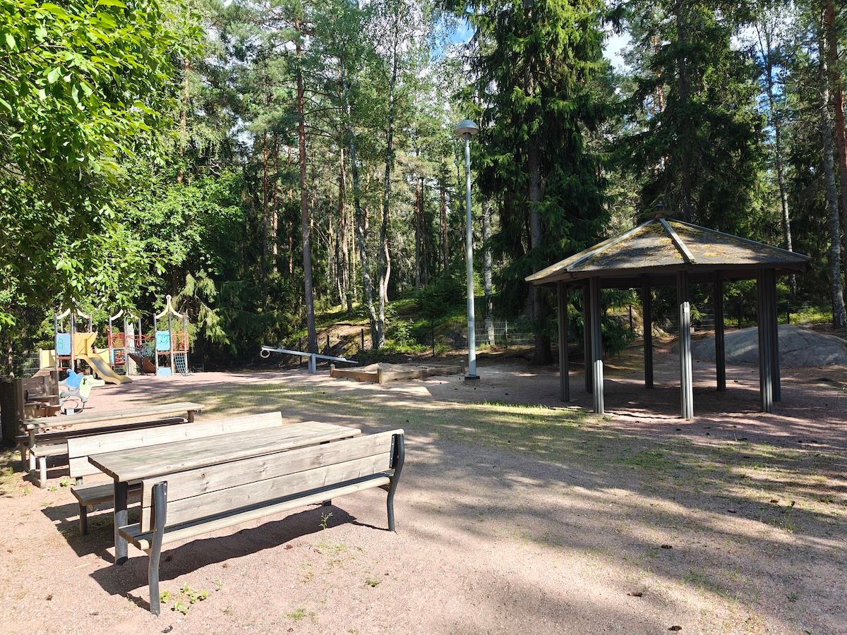 Yleiskuva leikkipaikasta, kiipeilyteline, penkit ja katos. Taustalla metsää.