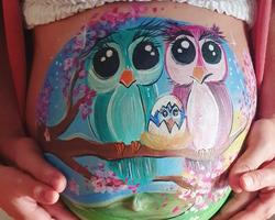 Diseño de bellypainting en la barriga de una mujer embarazada en el que aparecen dos búhos con su pollito saliendo del cascarón en un cerezo y durante un amanecer