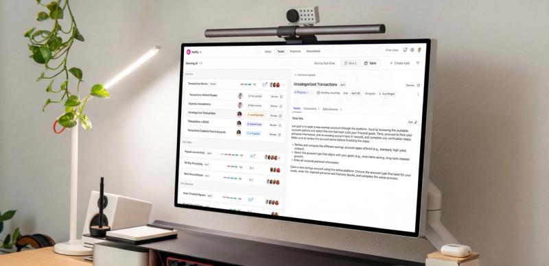 Desk setup with a monitor showing a task management dashboard, flanked by a webcam bar, LED lamp, and a small potted plant.