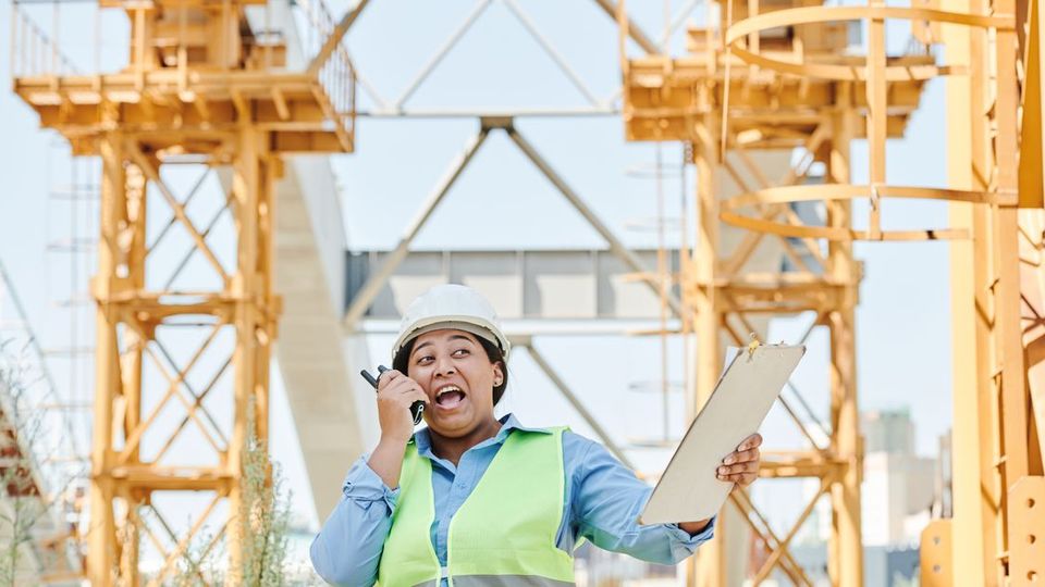 A site engineer in a hi-vis vest and hard hat talking on a two-way radio in front of yellow gantry cranes