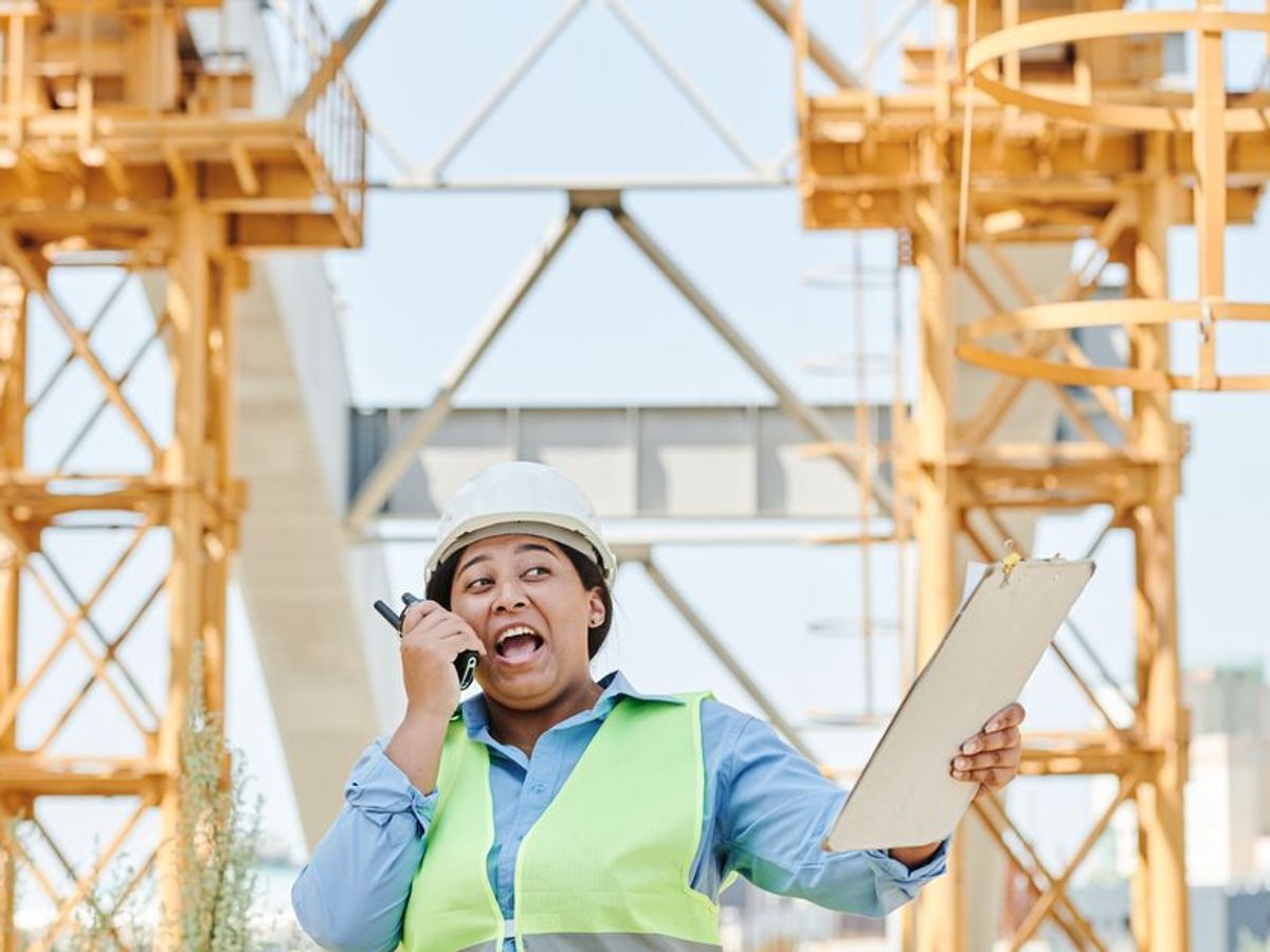 A site engineer in a hi-vis vest and hard hat talking on a two-way radio in front of yellow gantry cranes