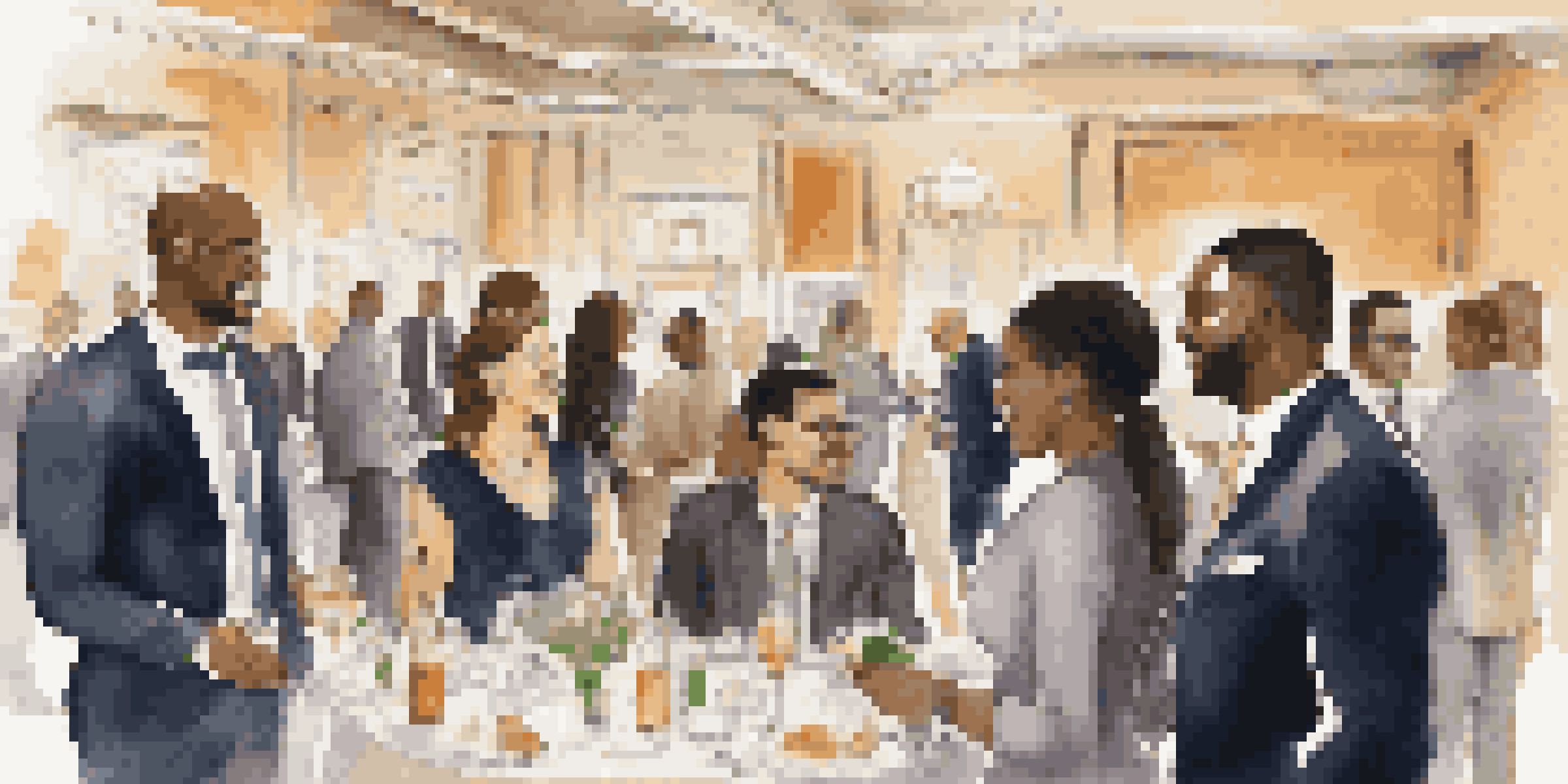 A diverse group of professionals in formal attire networking at an event, engaged in conversation with a table of business cards in the background.