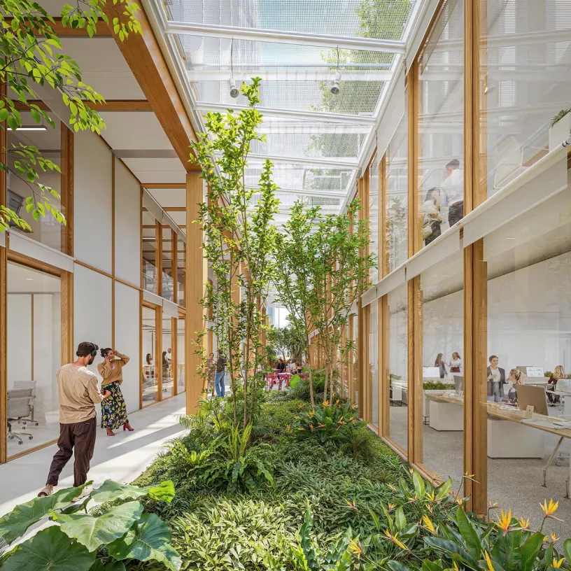 A bright, modern office with a central lush indoor garden surrounded by glass-walled workspaces on two levels, with people working and socializing.