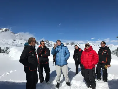 Seven people in winter jackets stand in deep snow with snow-covered mountains and a clear blue sky in the background.