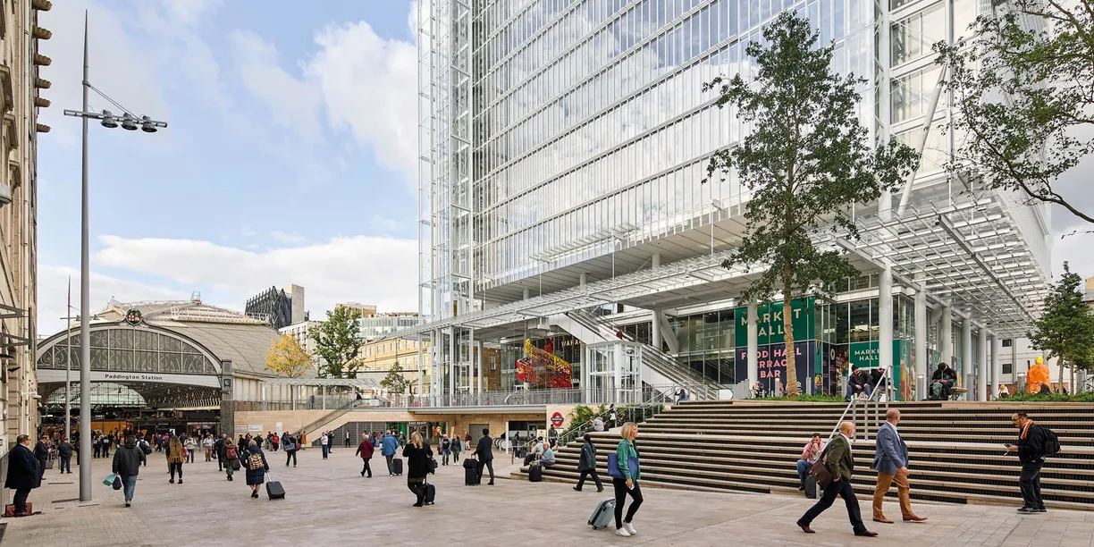 A busy urban plaza with a modern glass skyscraper, an old train station, and many people walking.