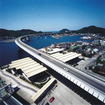 Aerial view of a curved bridge over a blue bay, connecting a coastal town nestled by green hills under a clear blue sky.