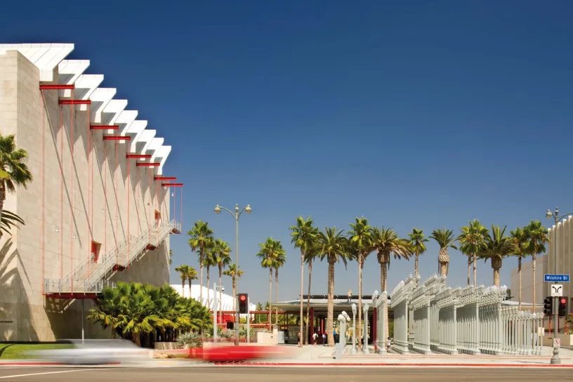 A red car is driving past a large building with palm trees in front of it