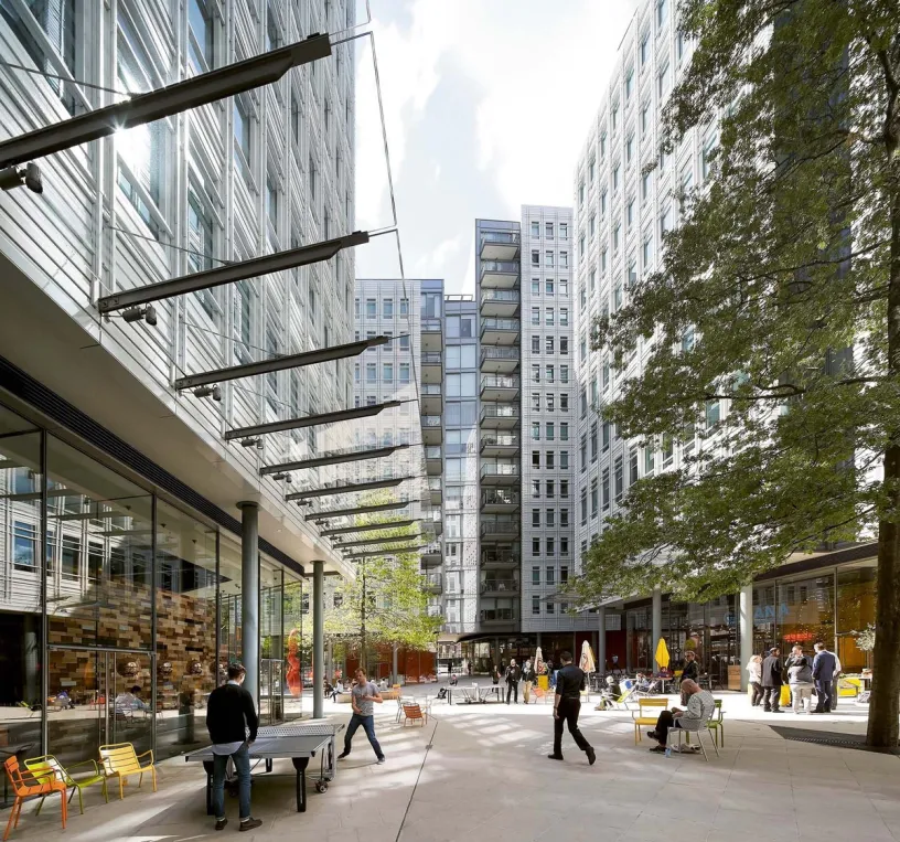 A modern urban plaza flanked by tall buildings, with people playing ping-pong and relaxing under trees.