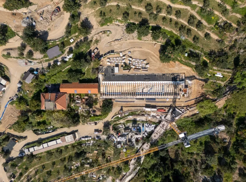 Aerial view of a construction site with a long building under construction, an older red-roofed building, a large crane, dirt roads, and terraced terrain.