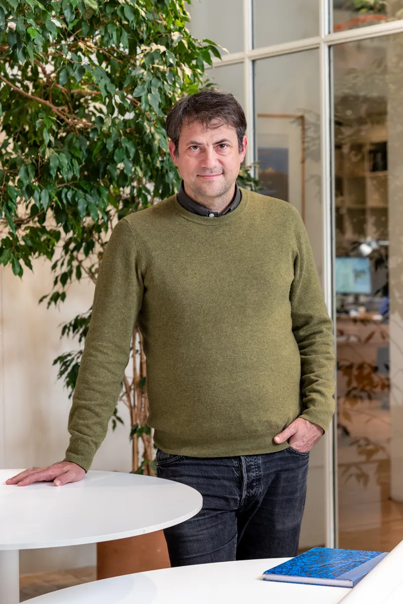 a man in a green sweater leans on a table with his hands in his pockets