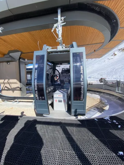 A gray ski gondola cabin with open doors and a white box inside, at a mountain loading station.