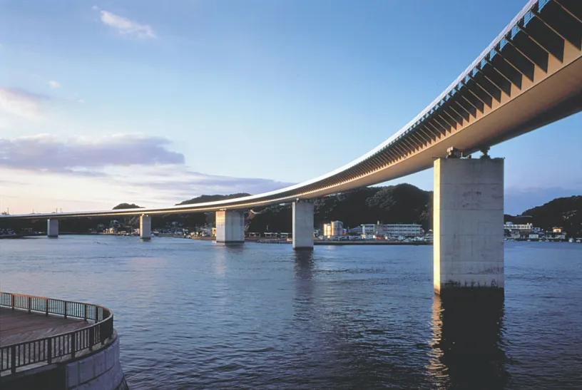 A long, curved bridge on concrete piers stretches across a body of water towards a distant town and hills.