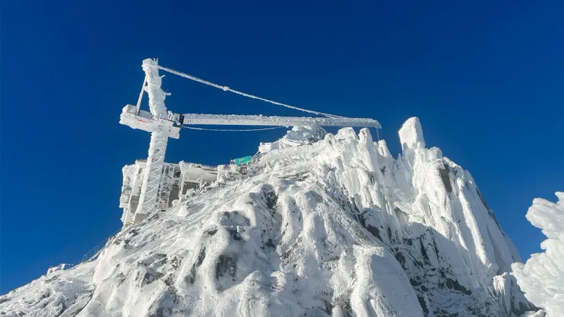 An ice-covered construction crane on a snowy mountain peak against a blue sky.