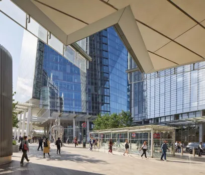 A bustling modern plaza with tall glass buildings, a metallic sculpture, and London Underground entrances on a sunny day.