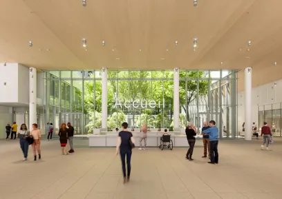 A bright, modern reception area with a wooden ceiling, a large glass wall overlooking trees, a counter labeled "Accueil," and several people.