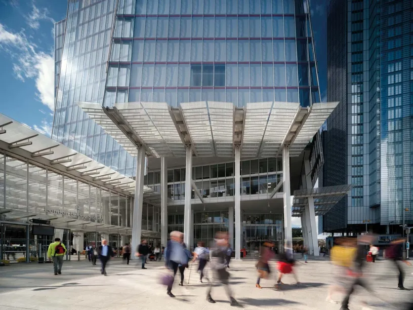A modern glass skyscraper with a large canopy entrance and blurred people walking on the plaza.