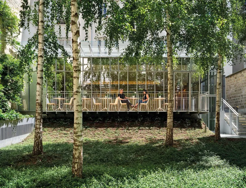 A couple of people are sitting at tables in front of a building surrounded by trees