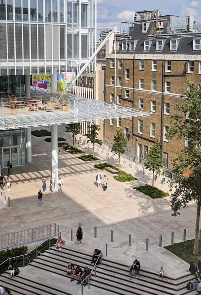 A vibrant modern public space with a large, swirling red and yellow abstract sculpture suspended overhead, people walking on a raised bridge and escalators, and Paddington Station in the background.