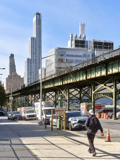 A person walks under an elevated train track on a city street lined with parked vehicles and tall buildings.