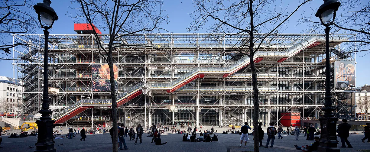 A large building with stairs leading up to it is surrounded by trees and people