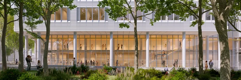 A modern building with a transparent ground floor showing people inside, framed by trees and green landscaping.