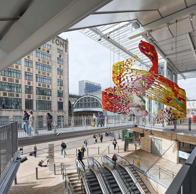 A vibrant modern public space with a large, swirling red and yellow abstract sculpture suspended overhead, people walking on a raised bridge and escalators, and Paddington Station in the background.