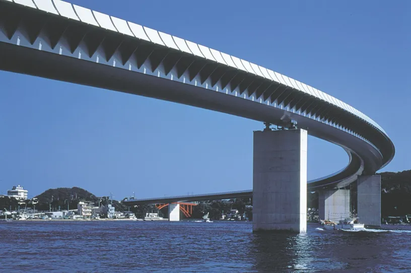 Modern curving bridge with saw-tooth panels on its underside, supported by concrete pillars over water.
