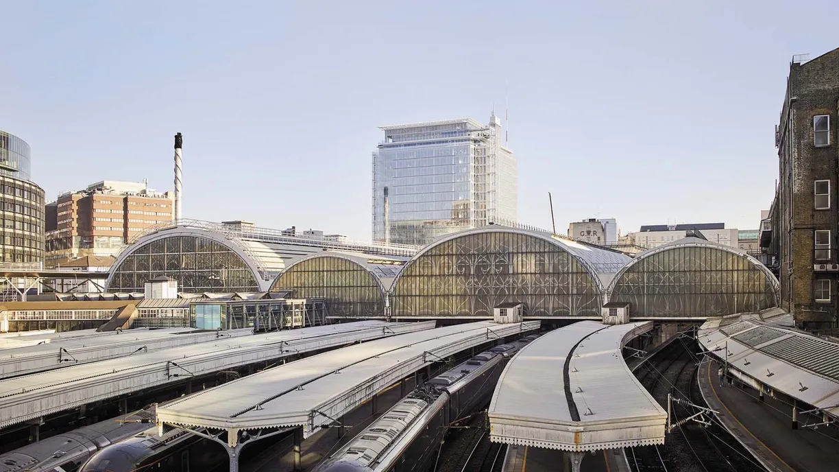 An elevated view of a train station with multiple platforms under arched glass roofs, surrounded by city buildings.
