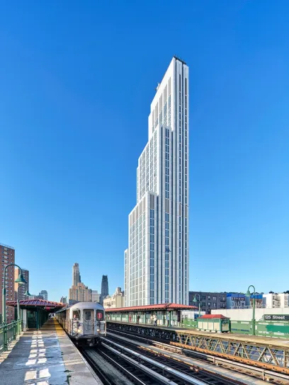 A tall, modern, stepped white skyscraper towers over an elevated subway station with a train.