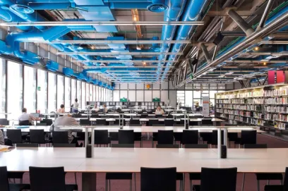 A large library with tables and chairs and blue pipes hanging from the ceiling