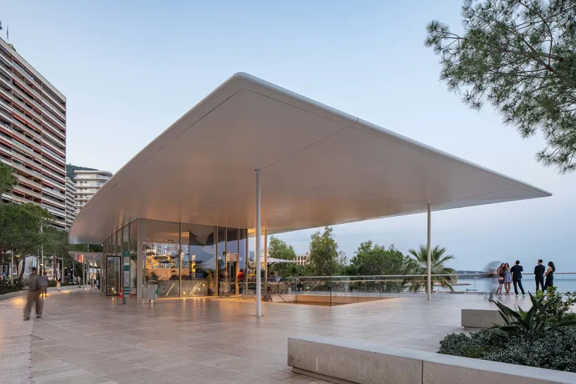 A group of people are standing under a large white roof in front of a building