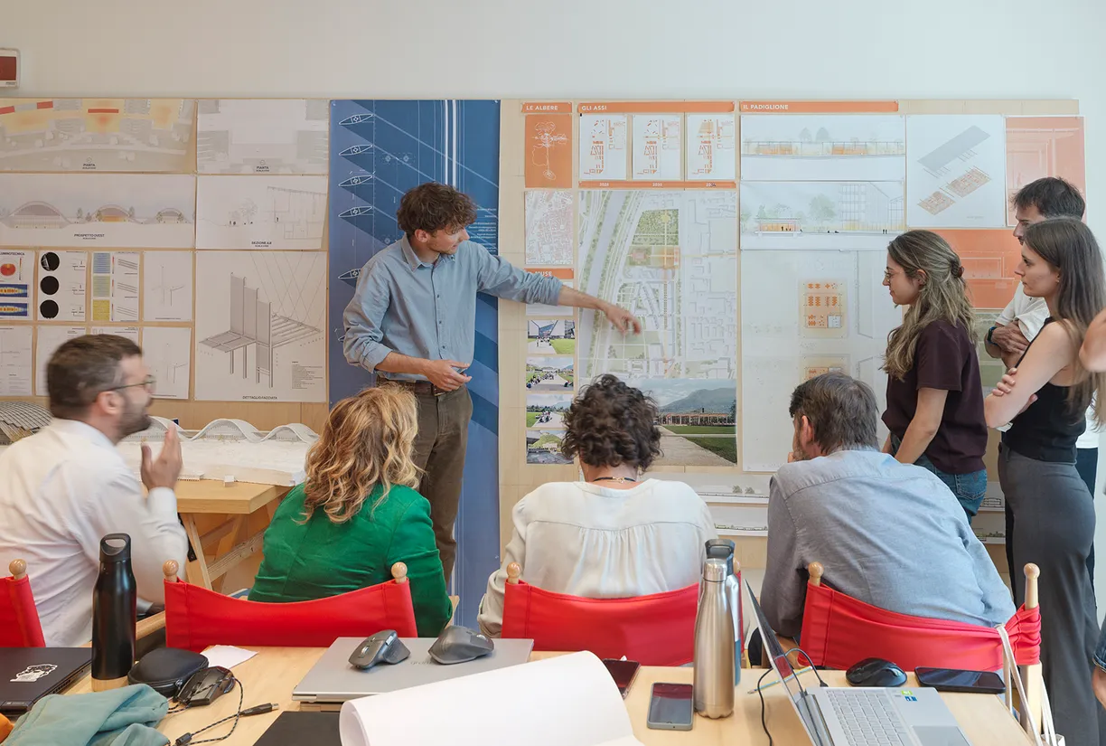 A man points to large architectural plans on a wall while presenting to a group of people in a bright room.