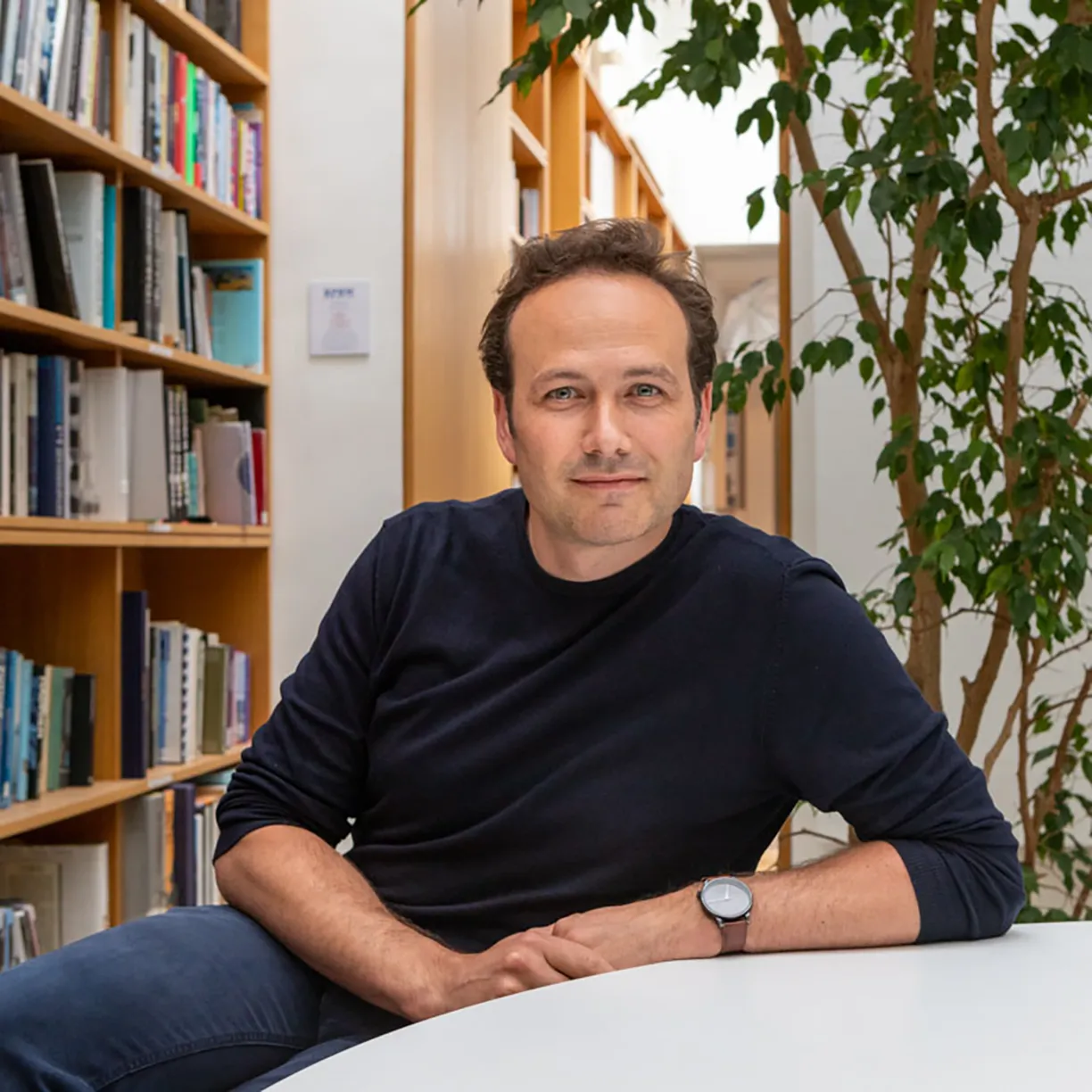 A man wearing a watch sits at a table in front of bookshelves