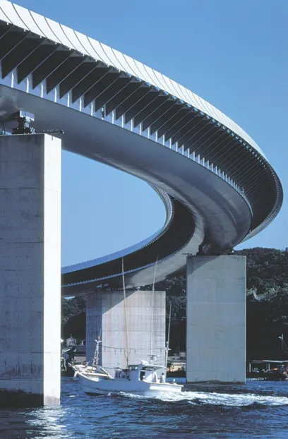 A curving bridge with a finned top is supported by concrete pillars over water, with a white boat passing beneath.