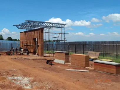 Construction workers building a structure with rammed earth walls and a metal roof frame on a dirt site.