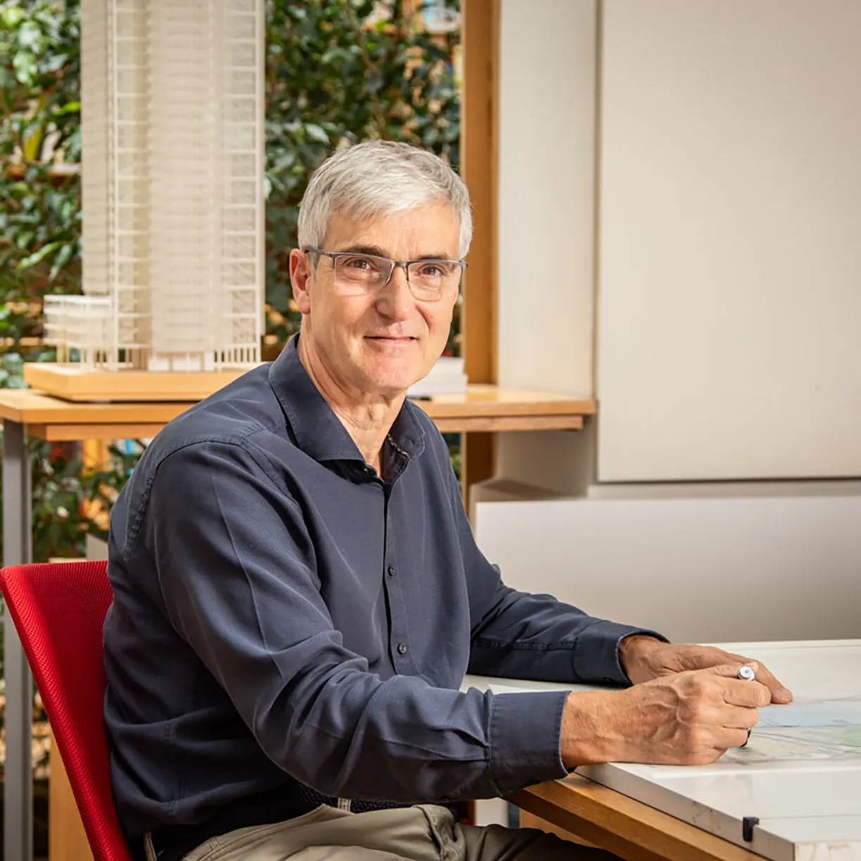 A man wearing glasses sits at a desk in front of a model of a building