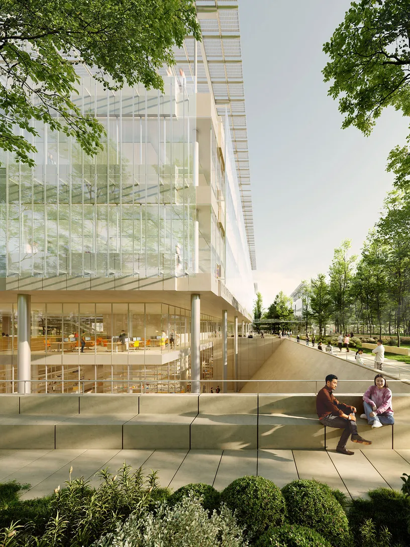 A bright, modern glass building surrounded by lush greenery, with two people sitting on an outdoor concrete bench.