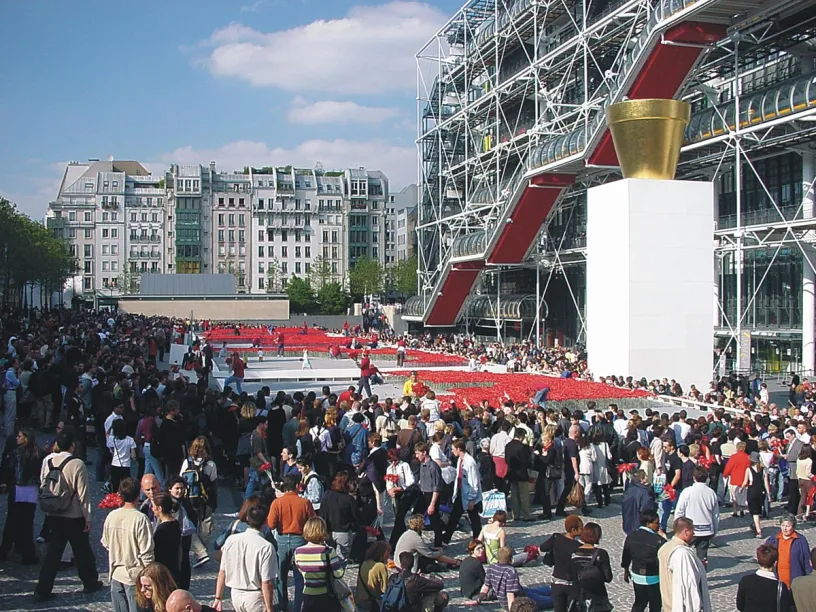 A busy plaza outside the Centre Pompidou, featuring its external red escalators, a large gold pot sculpture on a white pedestal, and an art installation of red poppies among a large crowd.