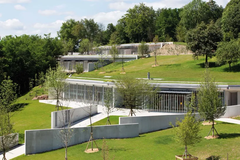 A large building is sitting on top of a lush green hill surrounded by trees